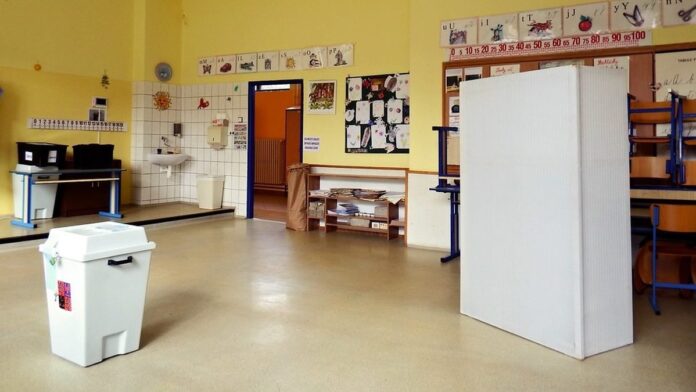 Volební místnost ve třídě základní školy s bílou volební urnou a plentou